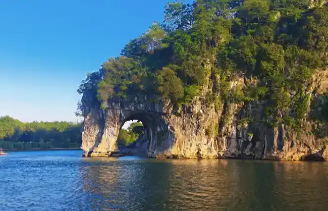 Rocky cliff with a circular natural arch carved into it, rising from the water with trees on top and a clear blue sky above.