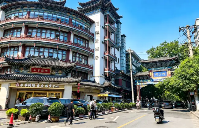 xiangzimiao-street | China Xian Tour: Private & Tailor-Made China Tours Busy street lined with ornate, curved-roof Chinese-style buildings, shopfronts on the ground floor, with parked cars, pedestrians, and a scooter under a bright blue sky.