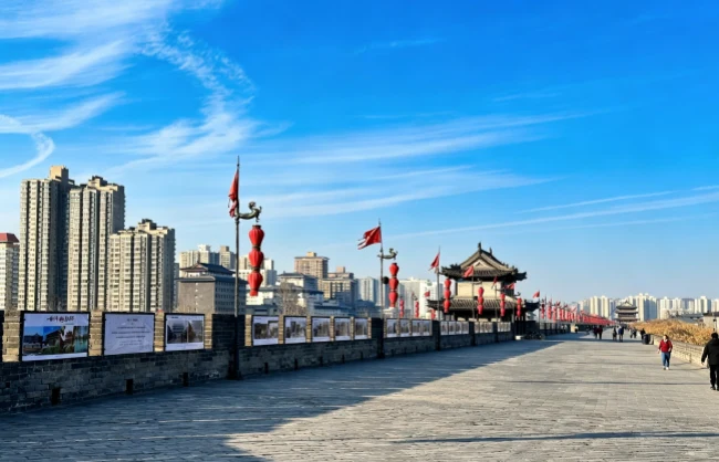 sunny-january-day-on-xian-city-wall-in-winter | China Xian Tour: Private & Tailor-Made China Tours Wide stone walkway along a historic city wall with red lanterns, a traditional gate, and modern high-rises in the background under a blue sky.