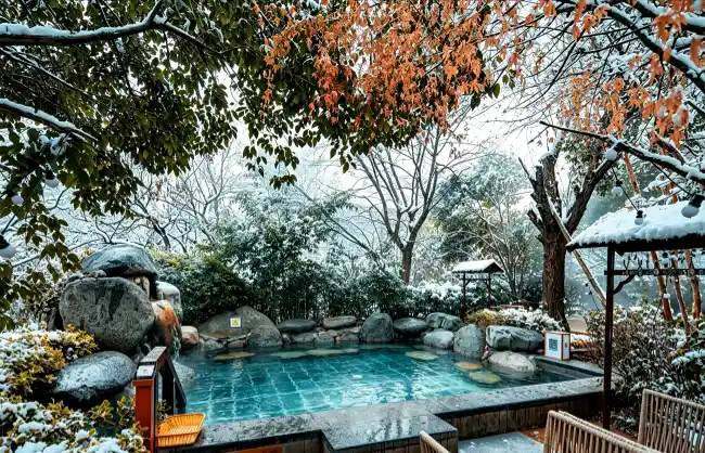 xian-hot-spring-in-january | China Xian Tour: Private & Tailor-Made China Tours Outdoor hot spring pool in a snow-covered garden, surrounded by rocks and trees with orange autumn leaves overhead.