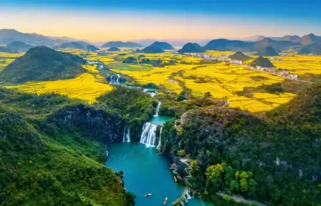 rapeseed-flowers-in-luoping-yunnan | China Xian Tour: Private & Tailor-Made China Tours Aerial view of bright yellow rice fields surrounding karst hills with a turquoise river and a foreground waterfall in the valley.