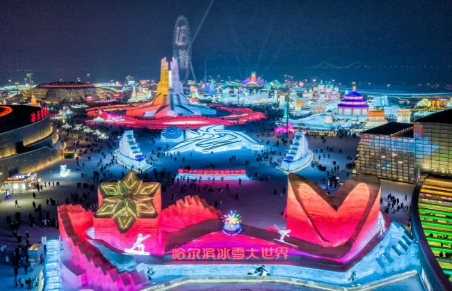 Nighttime view of a large illuminated ice and snow festival with neon-colored sculptures, winding paths, and crowds of visitors across the icy grounds.