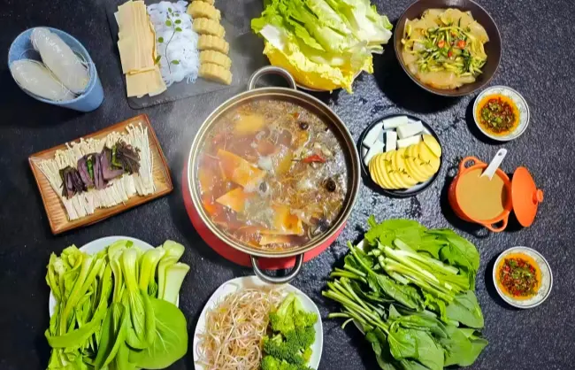 vegetable for hot pot | China Xian Tour: Private & Tailor-Made China Tours Top-down view of a pot of simmering soup surrounded by fresh vegetables, noodles, and dipping sauces.