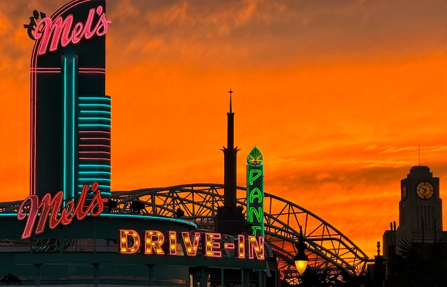 mels-drive-in | China Xian Tour: Private & Tailor-Made China Tours Neon Mel's Drive-In signs with DRIVE-IN letters, silhouetted roller-coaster/bridge under an orange sunset.