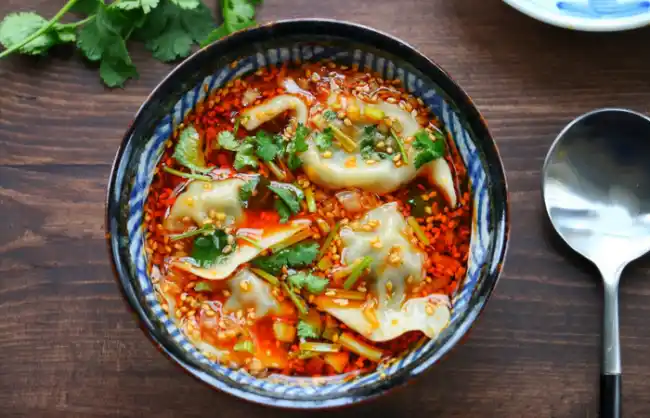 dumplings | China Xian Tour: Private & Tailor-Made China Tours Dumplings in a spicy red chili oil with cilantro garnish in a blue-rimmed bowl on a wooden table.