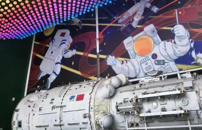 astronaut-experience-china-science-and-technology-museum - China Xian Tour: Private & Tailor-Made China Tours White space capsule replica on a display stand, with a vibrant mural of astronauts and space in the background; a Chinese flag is visible on the capsule.