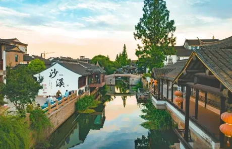 Canal lined with traditional Chinese buildings and lanterns, a wooden bridge crossing, and calm water reflecting the sky, trees, and architecture; a white wall with Chinese characters on the left.