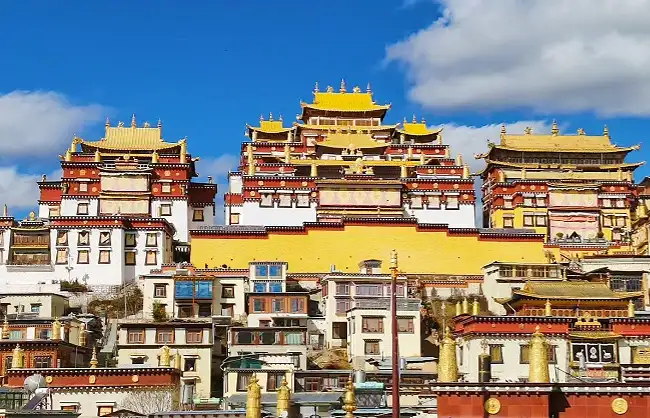 Tibetan monastery complex with yellow-tiled roofs perched on a hillside under a blue sky, white and red walls visible in front.