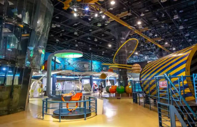 xiamen-science-technology-museum | China Xian Tour: Private & Tailor-Made China Tours Colorful indoor science museum scene with interactive exhibits, bright yellow walkways, and a striped tunnel structure in the background.