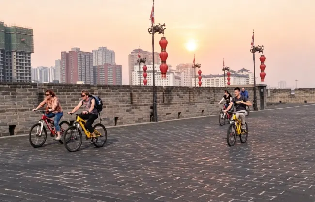city-wall-cycling-sunset | China Xian Tour: Private & Tailor-Made China Tours Four people ride bicycles along a brick city wall at sunset, with red lanterns lining the path and a distant skyline behind them.