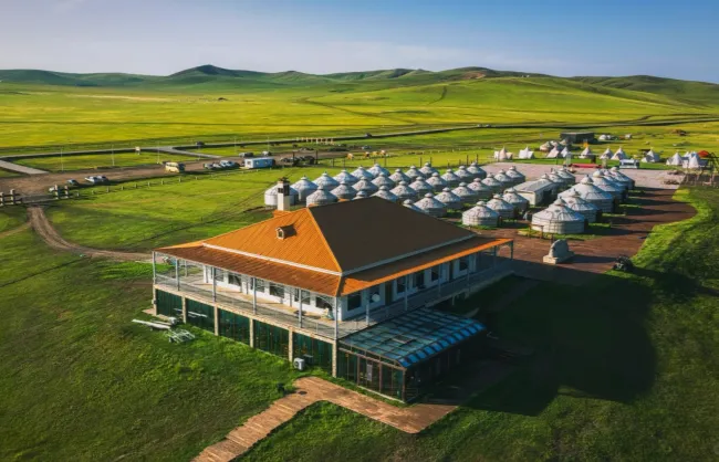Lodge with orange roof beside a field of white yurts on green grass under a blue sky.