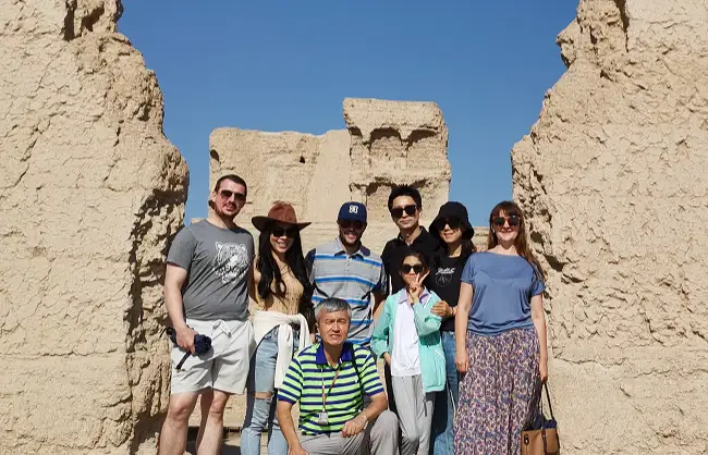 Group of eight people posing together in front of tall beige rock formations on a sunny day. Some wear hats and sunglasses while smiling for the photo, suggesting a sightseeing outing.