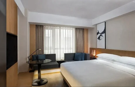 Hotel room with a large white-bed, nightstand lamp, and wall art; seating area with a round table and sofa by a window with sheer curtains