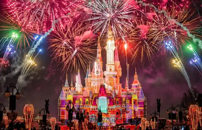 Castle lit up with colorful lights at night as fireworks explode overhead in a festive display.