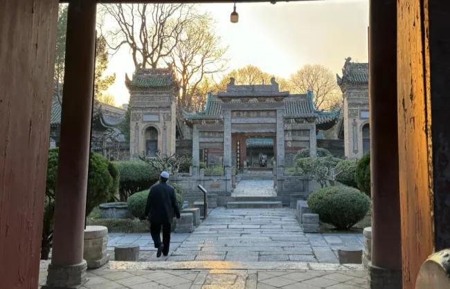 Man in a dark suit walking along a stone path toward a traditional Chinese temple gate, framed by arches and trimmed shrubs at sunset.